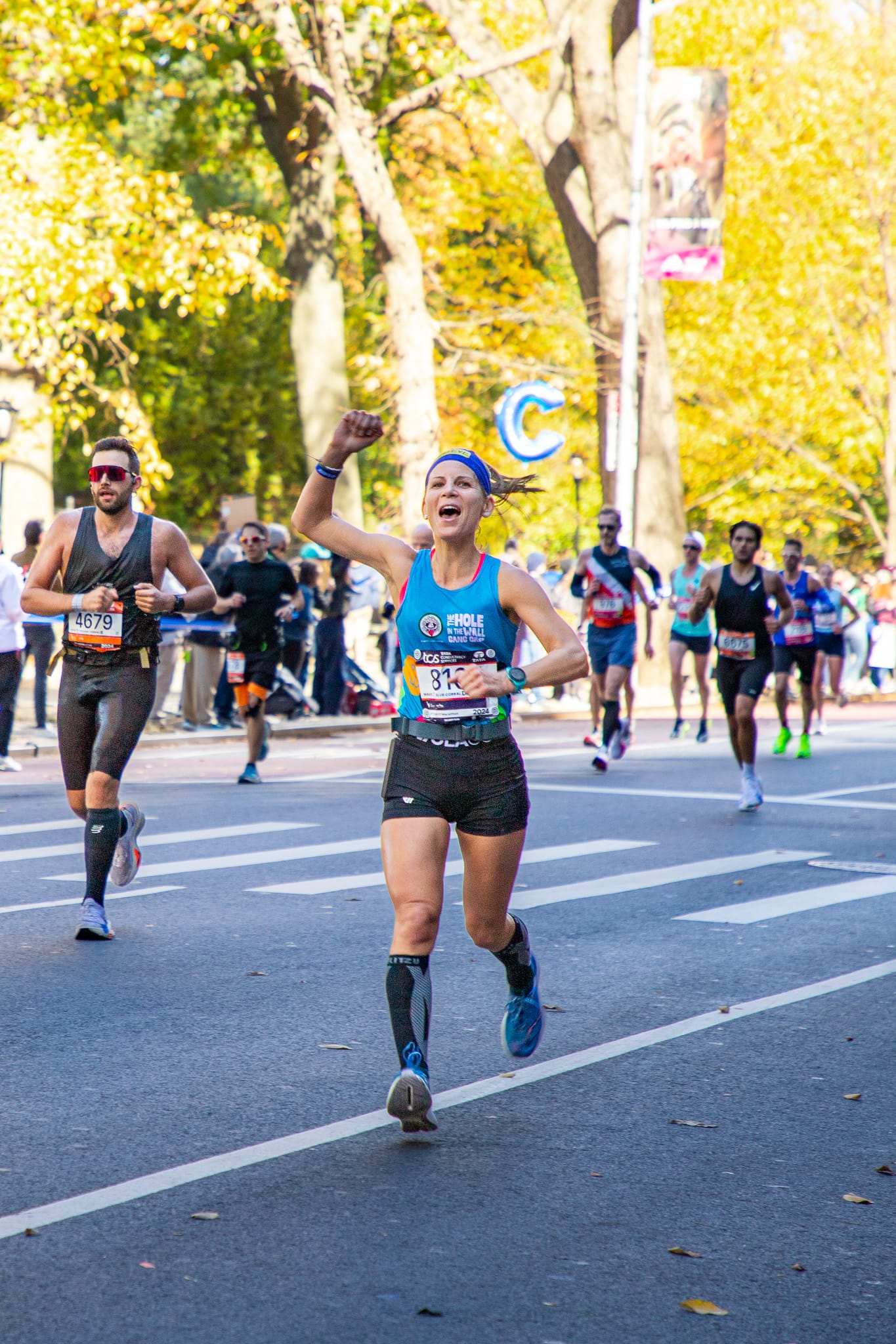 A smiling runner raises a fist in celebration, wearing a blue tank top and headband, as she nears the finish of a marathon. Other runners and spectators line the sunlit street with autumn trees.
