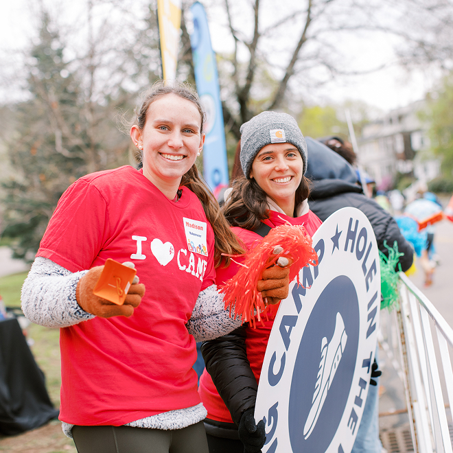 Two women in red shirts and winter hats smile at a race event. One holds a cowbell, the other holds pom-poms, and they stand by a large circular sign. Trees and race participants are visible in the background.