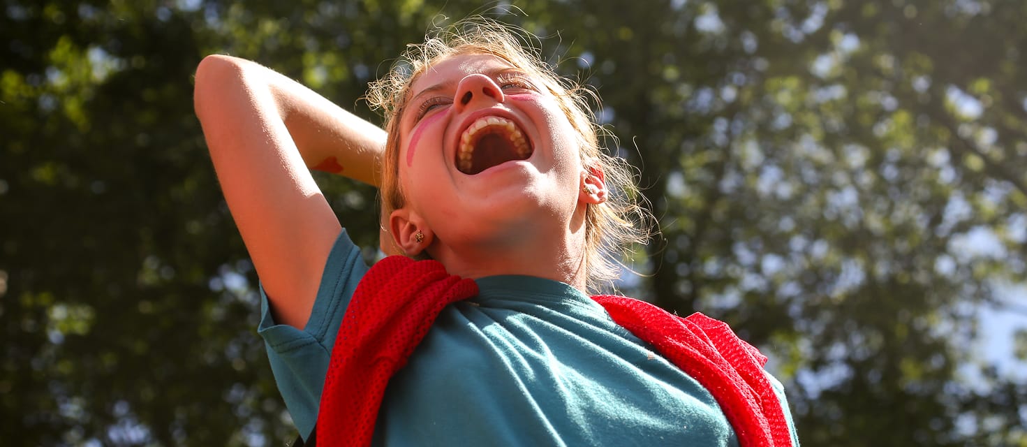 A child outdoors, laughing joyfully with head tilted back, one hand behind their head. They wear a blue shirt and a red sweater draped over their shoulders, with sunlight and green trees in the background.