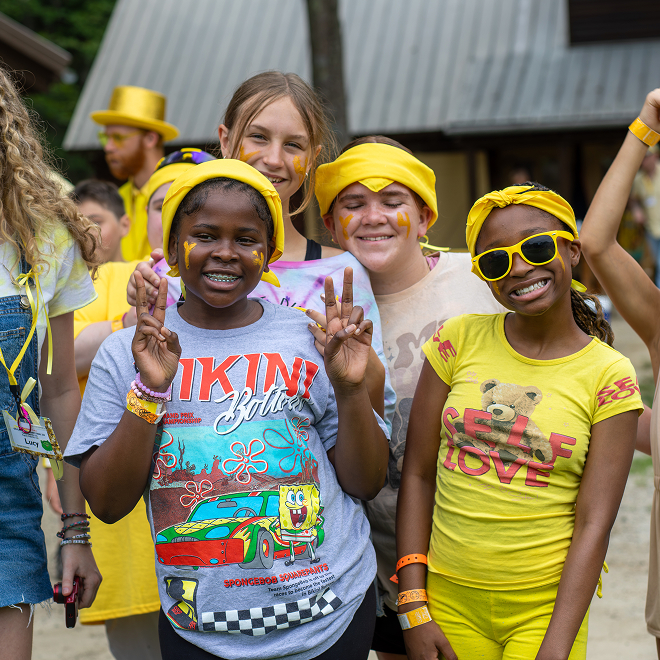 Four smiling children pose outdoors at camp, wearing yellow accessories and face paint. One wears a SpongeBob shirt, another wears yellow sunglasses and a Self Love shirt. Other campers in yellow are visible in the background.