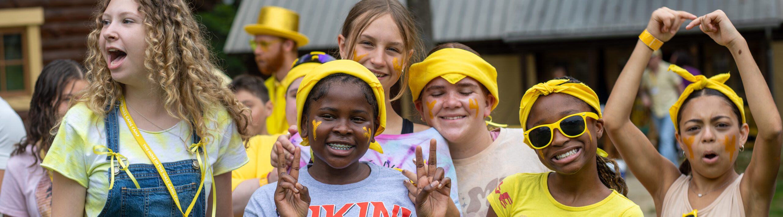 A group of smiling kids at camp, some wearing yellow bandanas and sunglasses, pose happily outdoors. One girl flashes peace signs, and another makes a heart shape with her hands.