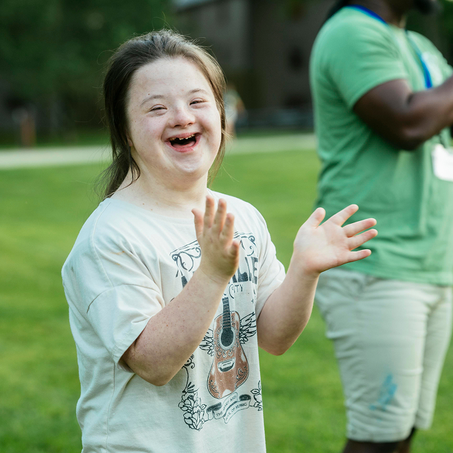 A person with Down syndrome smiles and claps outdoors, wearing a T-shirt with a guitar design. Another person in a green shirt stands nearby, blurred in the background.