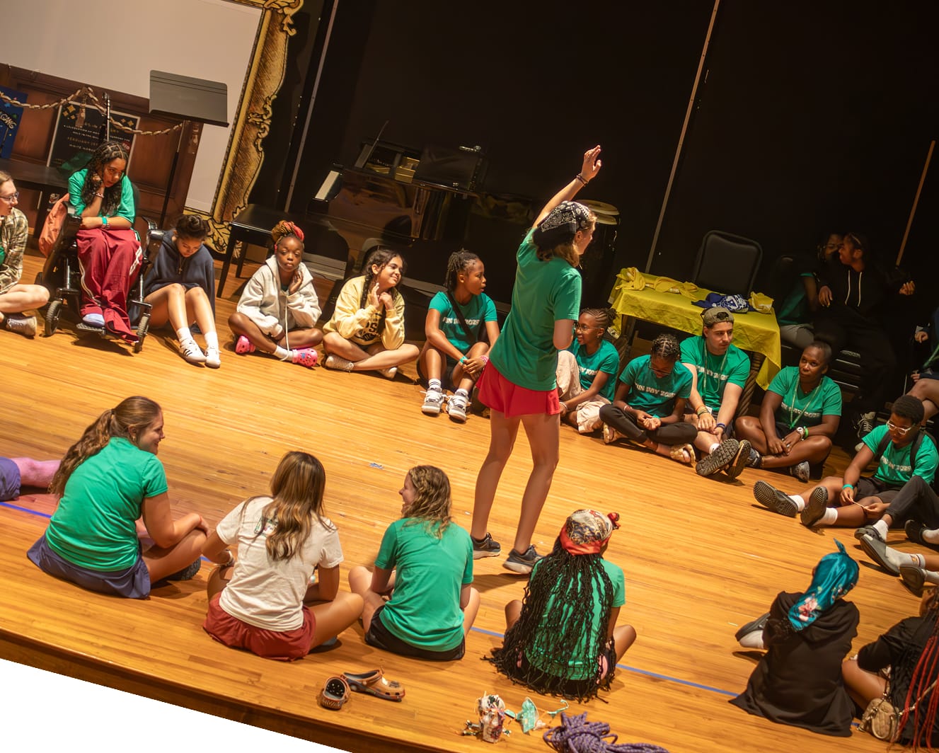 A group of children sit in a large circle on a stage, listening to a person standing in the center raising their hand. Most kids wear green shirts; musical instruments are visible in the background.