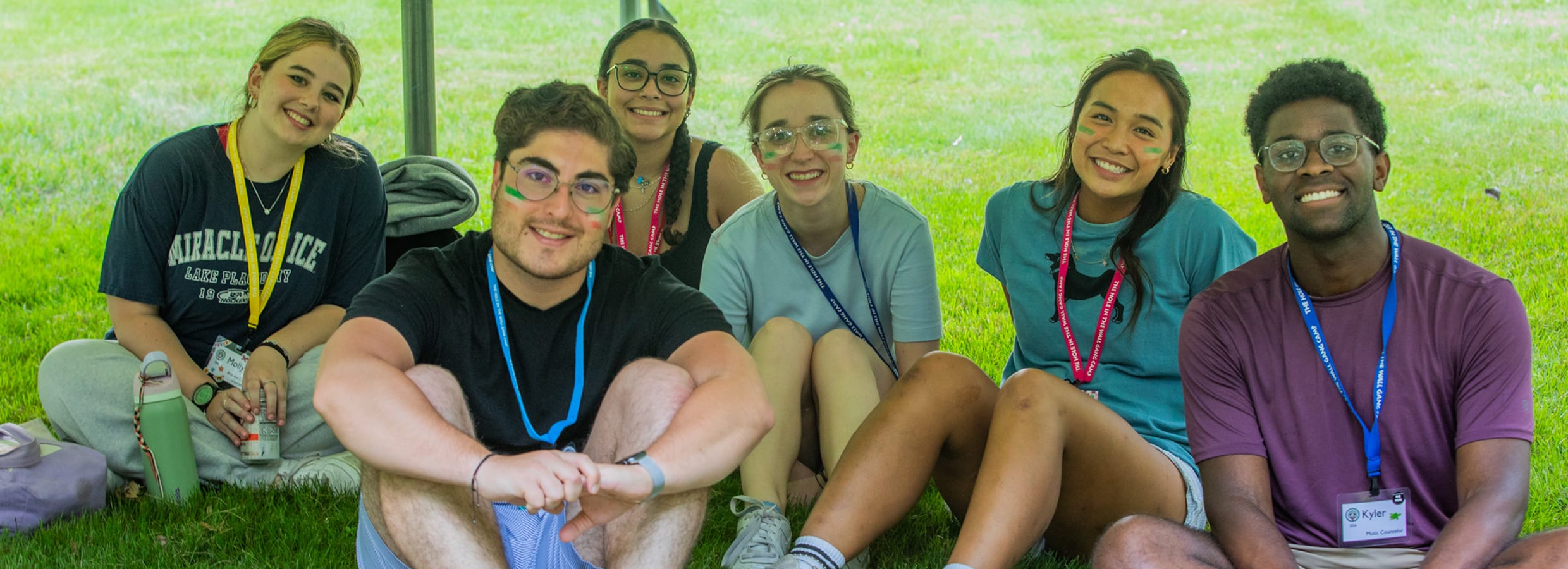 Six young adults sit together on grass under a canopy, smiling at the camera. They wear casual clothes and lanyards, suggesting a friendly or group event outdoors. Trees and green lawn are visible in the background.