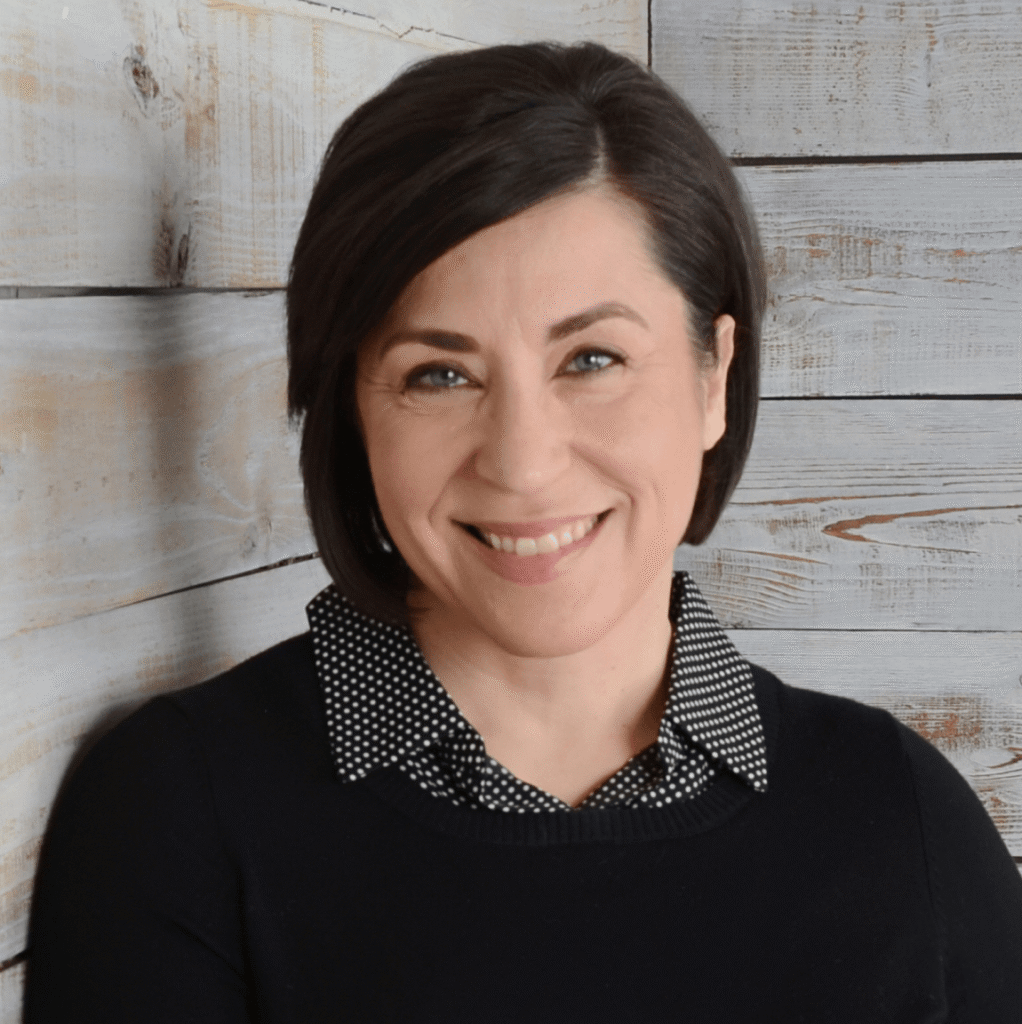 A woman with short dark hair smiles at the camera, wearing a black top with a black-and-white polka dot collar, standing in front of a light wooden plank background.
