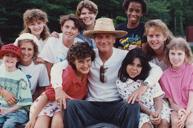 A group of smiling people, including children and adults, pose together outdoors. The man in the center wears a straw hat and sunglasses around his neck. Trees are visible in the background.