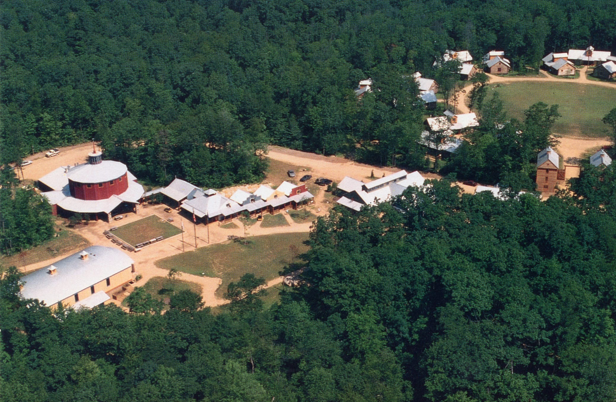 Aerial view of a rural camp with several cabins, a circular red building, open fields, and dirt paths surrounded by dense forest.