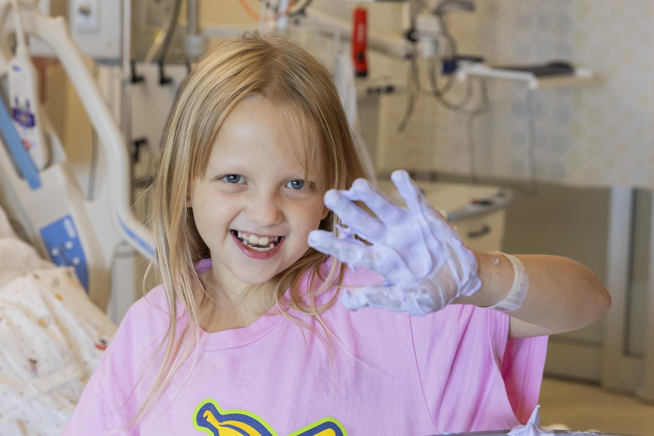 A smiling young girl in a hospital room shows her hand covered in purple paint while wearing a pink shirt. Medical equipment and a bed are visible in the background.