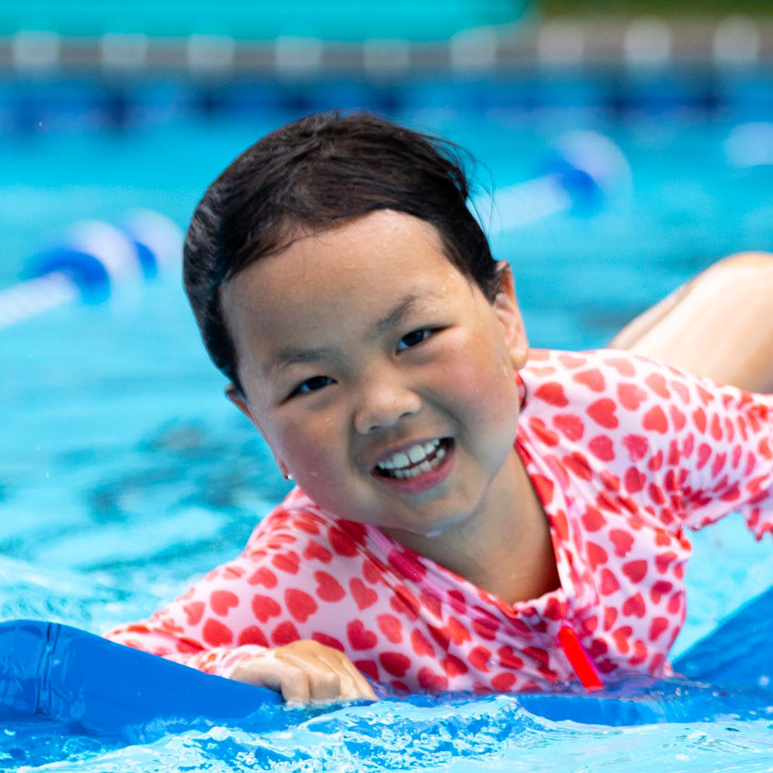 Smiling child in a heart-patterned swimsuit rides a blue pool float in a swimming pool, with water and pool lane markers visible in the background.