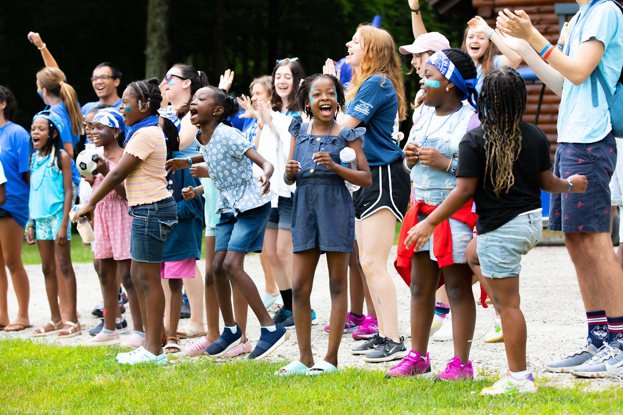 A diverse group of children and adults stand outdoors, smiling and cheering together. Many wear blue clothing and headbands, and some have their arms raised in excitement. Trees and a wooden building are in the background.