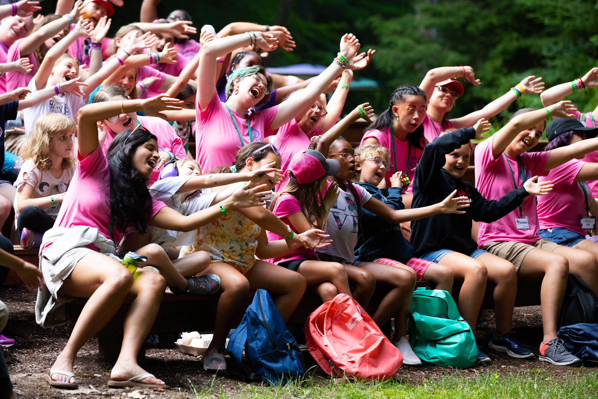 A group of kids and teens in pink shirts sit outdoors on benches, laughing and waving their arms enthusiastically in the same direction, with colorful backpacks at their feet.