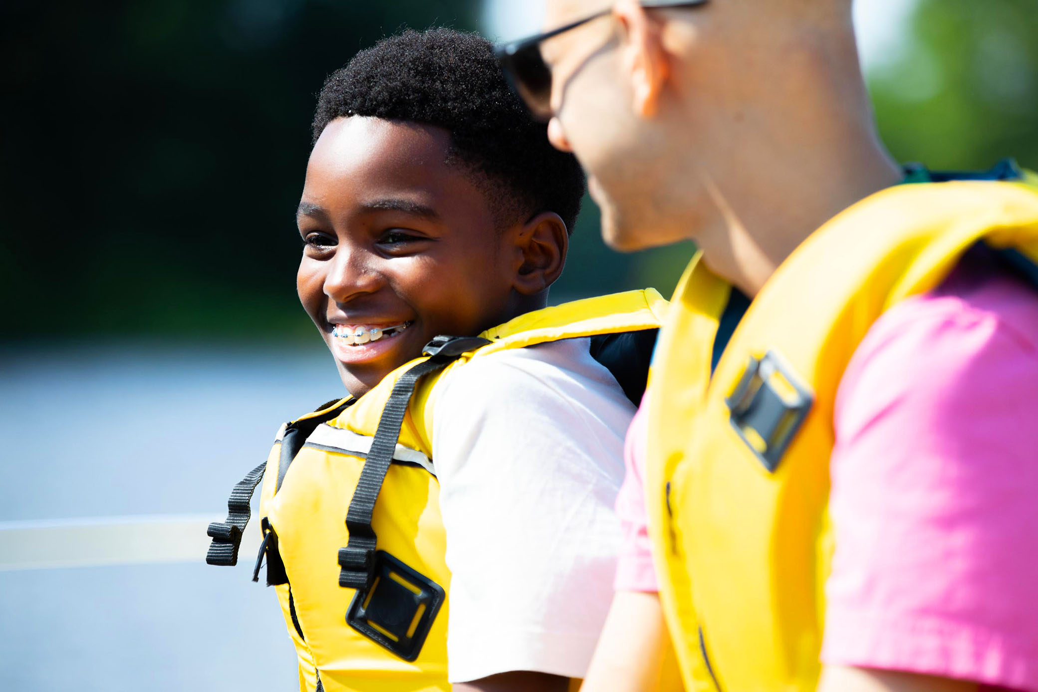 A smiling boy wearing a yellow life jacket sits next to an adult in a yellow life jacket and pink shirt, both enjoying a sunny day near the water.