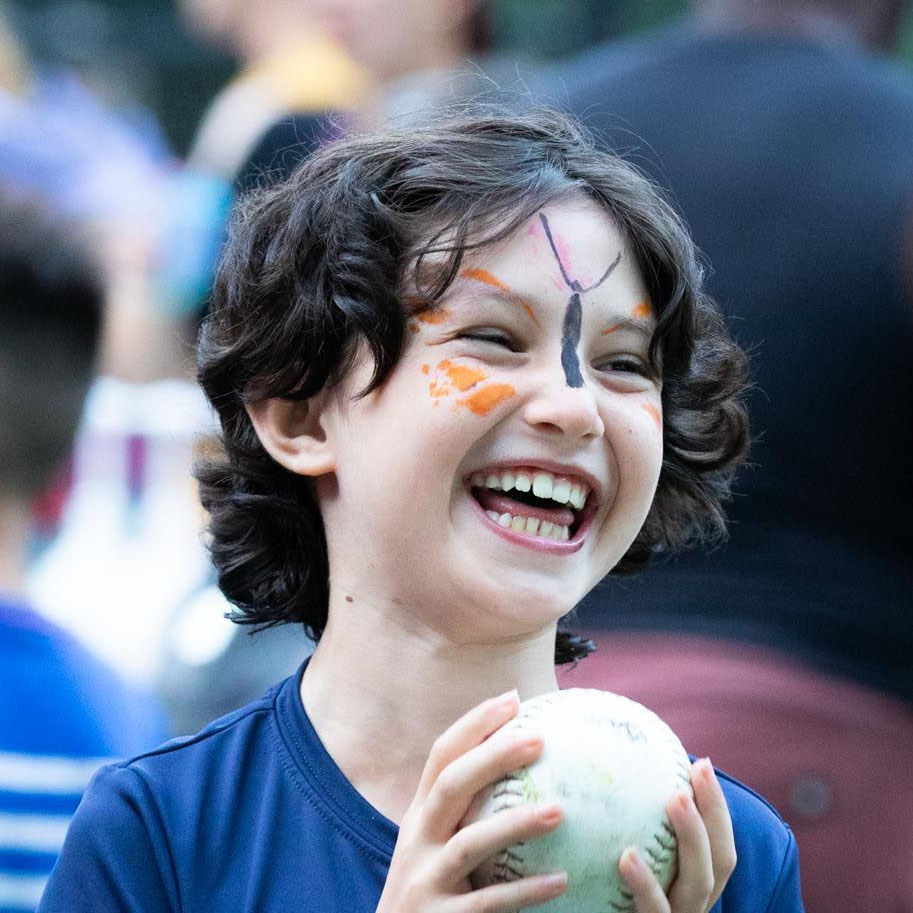 A child with curly hair and butterfly face paint smiles brightly while holding a baseball. The background is blurred with people, focusing attention on the childs joyful expression.