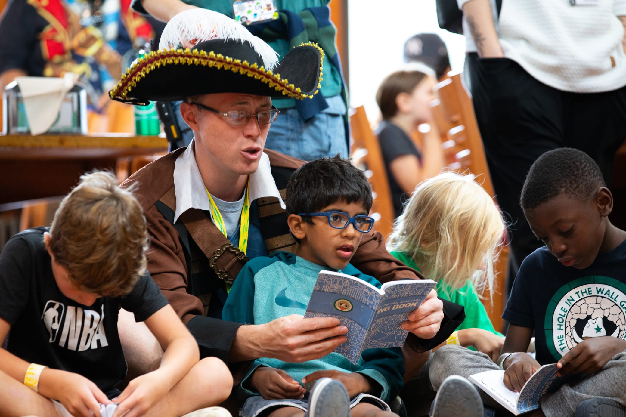 An adult in a pirate costume reads a book with a group of young children sitting on the floor. The children are focused on their own books. Other people are in the background, suggesting a group activity or event.