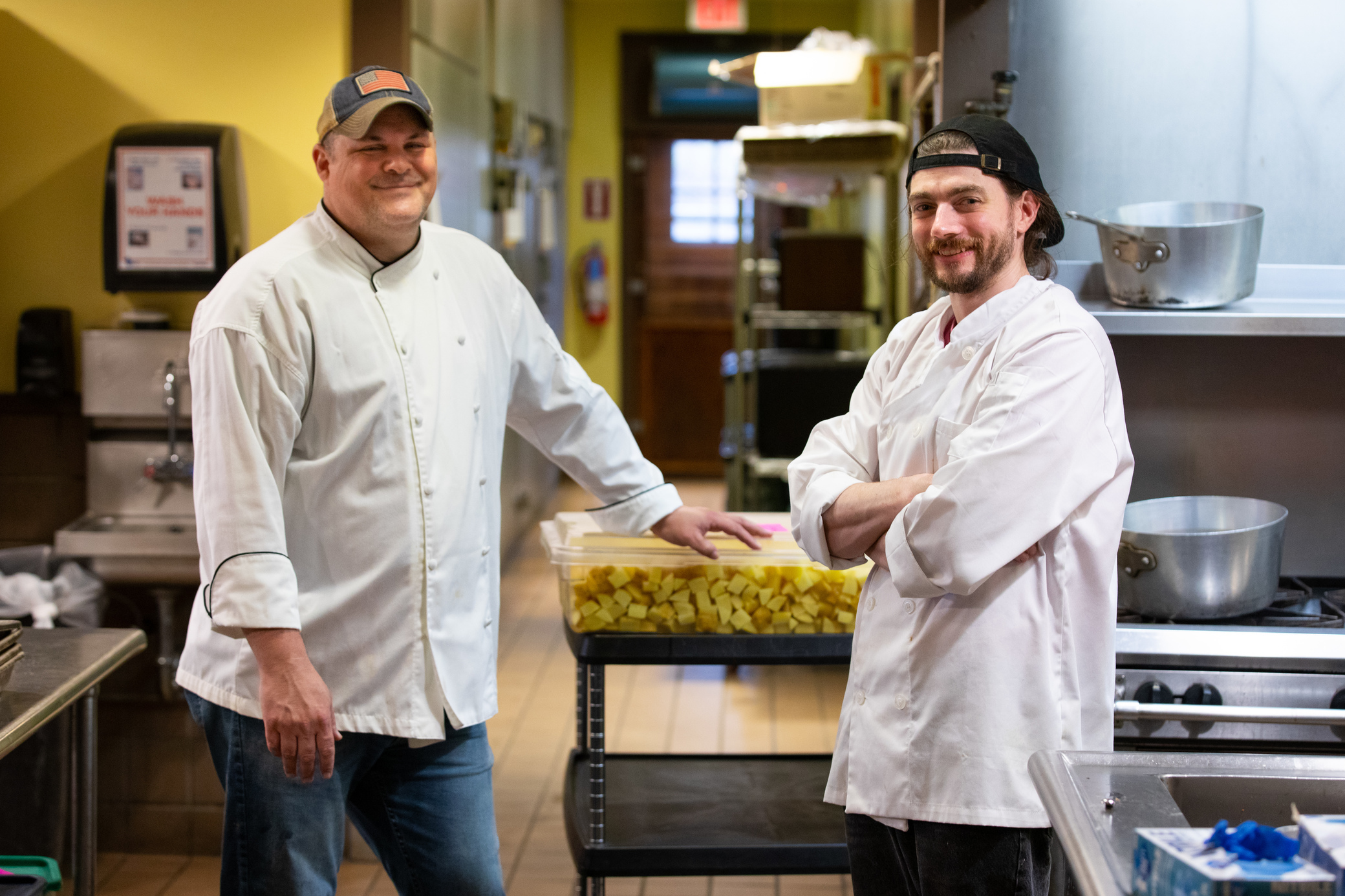 Two chefs in white jackets stand in a commercial kitchen, smiling at the camera. One stands with arms crossed, the other with a hand on a container of chopped potatoes. Stainless steel equipment surrounds them.