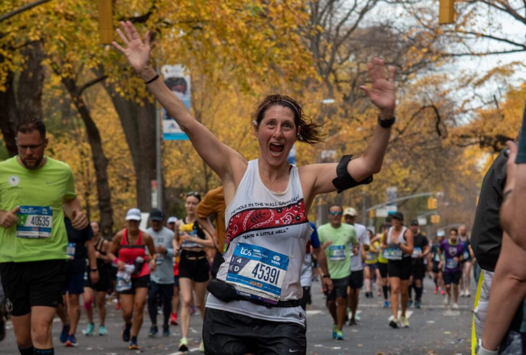 A smiling runner with bib number 45391 raises both arms in celebration during a marathon on a tree-lined street filled with other runners on an autumn day.