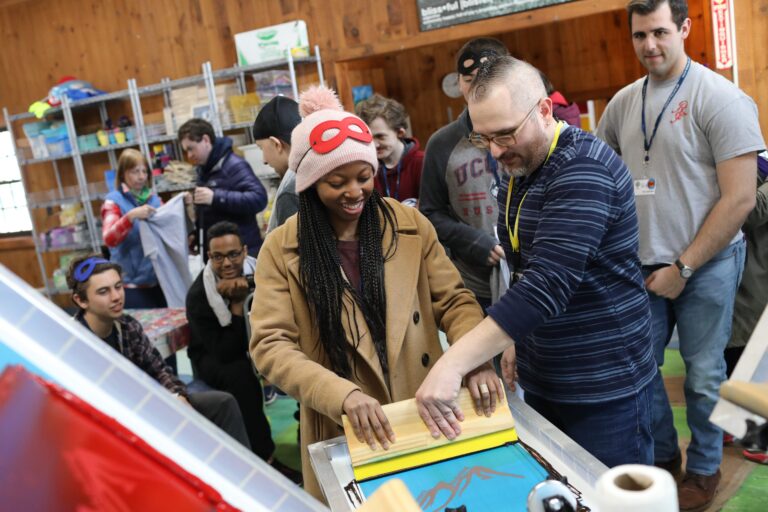A group of people watch as a smiling woman and a man work together on a screen printing project indoors. The woman wears a tan coat and pink hat, while the man guides her hands as they print on blue fabric.