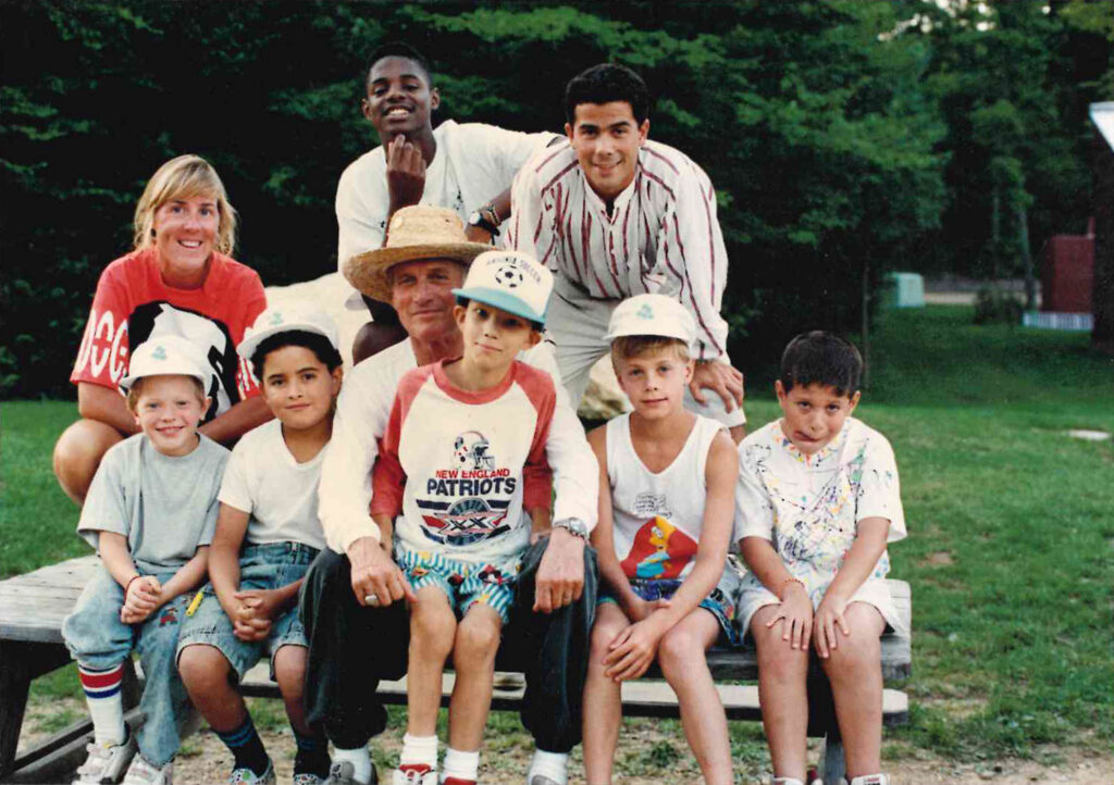 A group of eight people, including children and adults, pose together outdoors on a bench with trees in the background. Some wear hats and casual summer clothes, and all are smiling or looking at the camera.