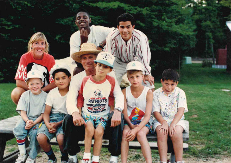 A group of eight people, including children and adults, pose together outdoors on a bench with trees in the background. Some wear hats and casual summer clothes, and all are smiling or looking at the camera.