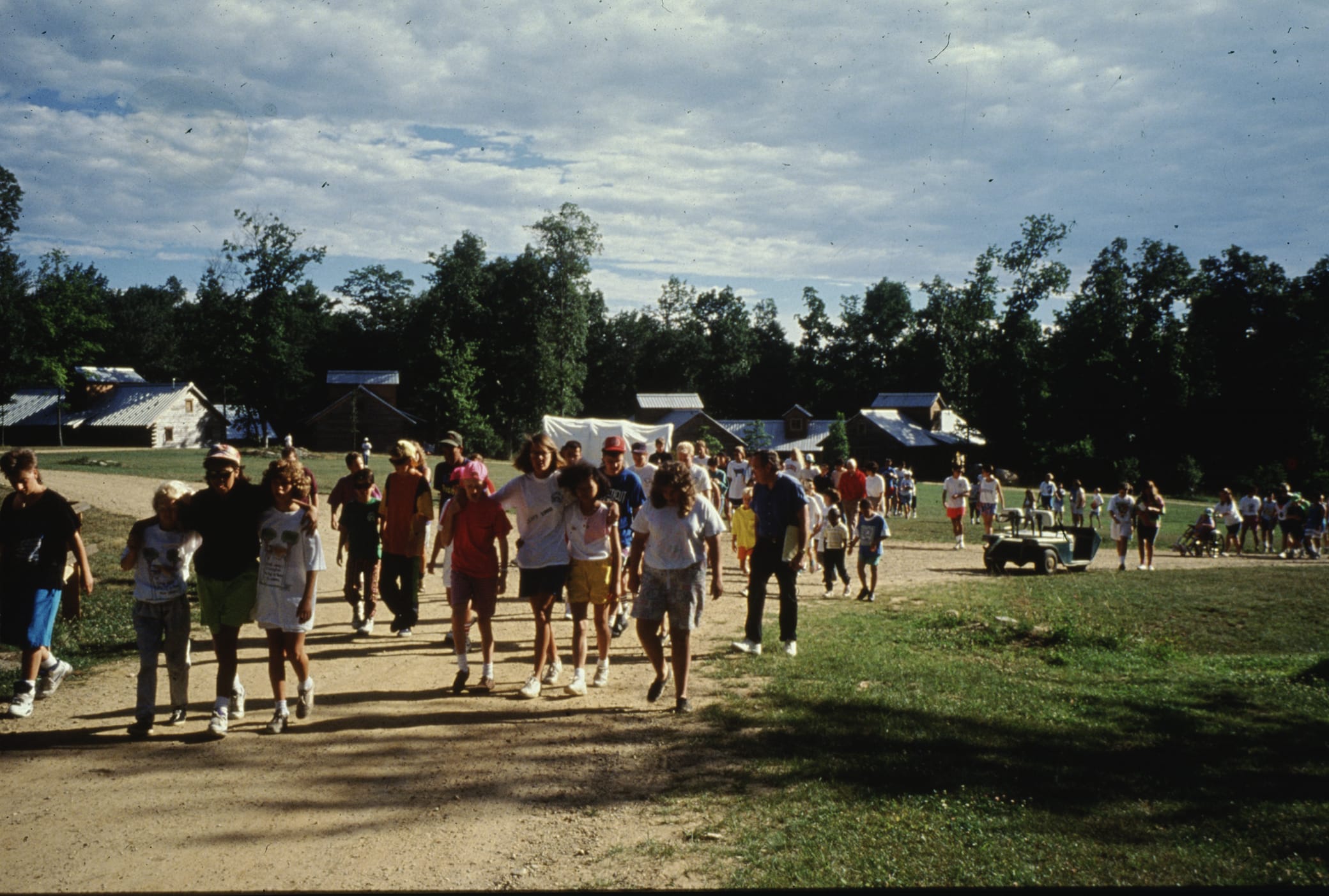 A large group of people, mainly children, walk along a dirt path at a camp with cabins and tents in the background, surrounded by trees under a partly cloudy sky.