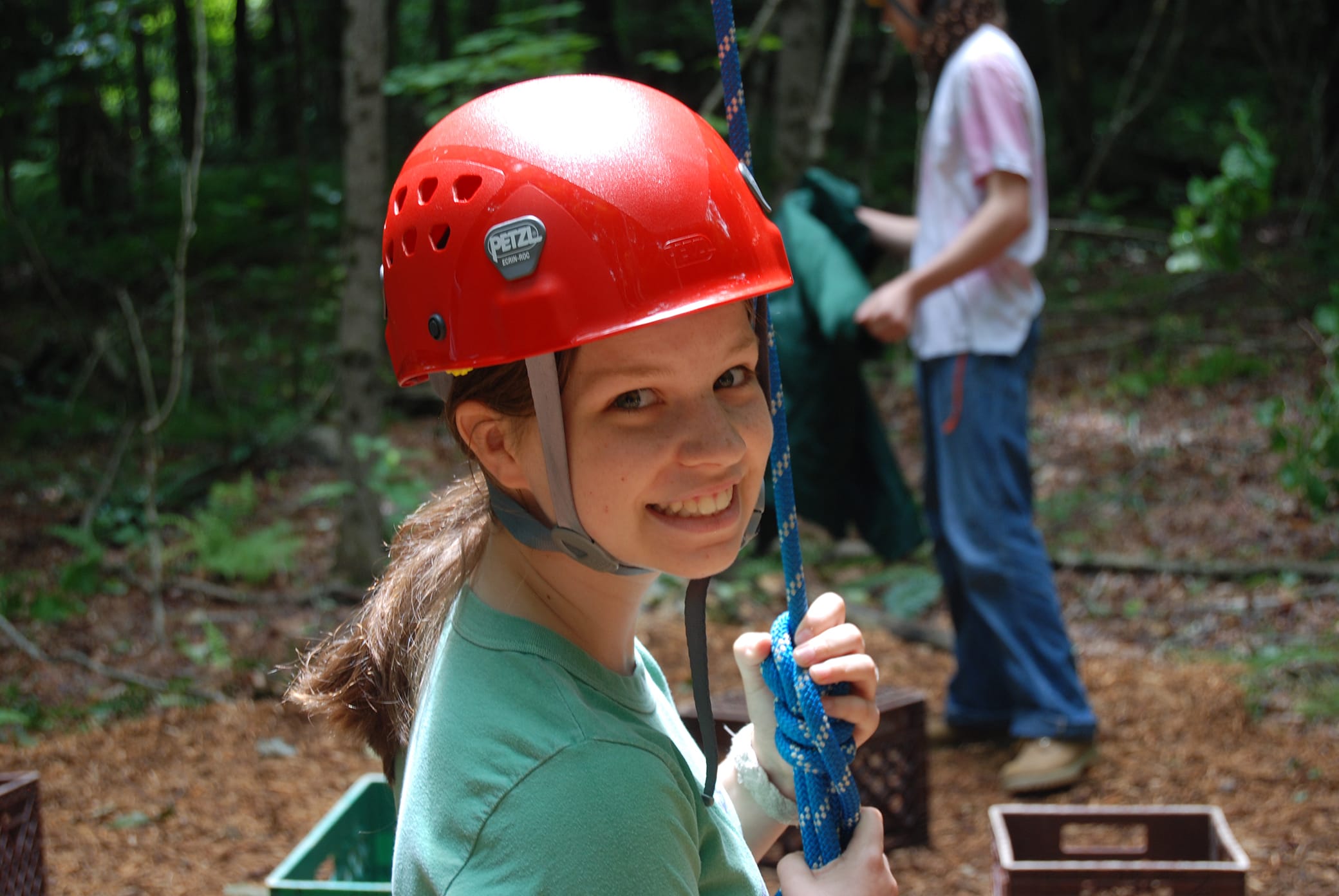 Smiling person in a red helmet holds a blue rope outdoors with trees and crates nearby; another person, blurred in the background, stands behind them.