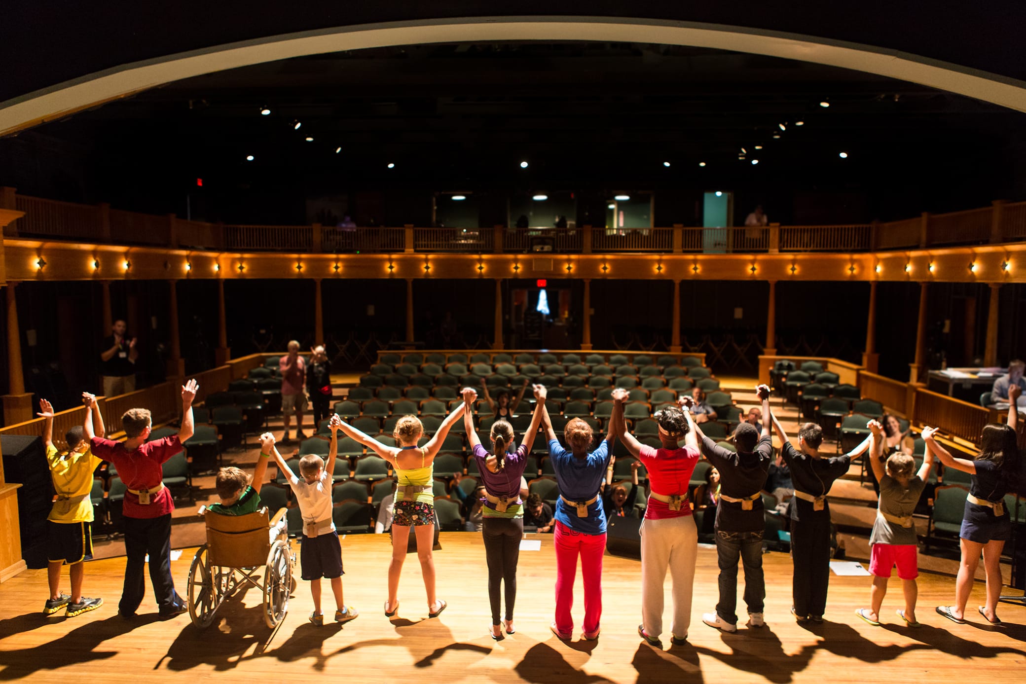 A diverse group of performers, including one in a wheelchair, stand onstage holding hands and raising their arms to an applauding audience in a warmly lit theater.