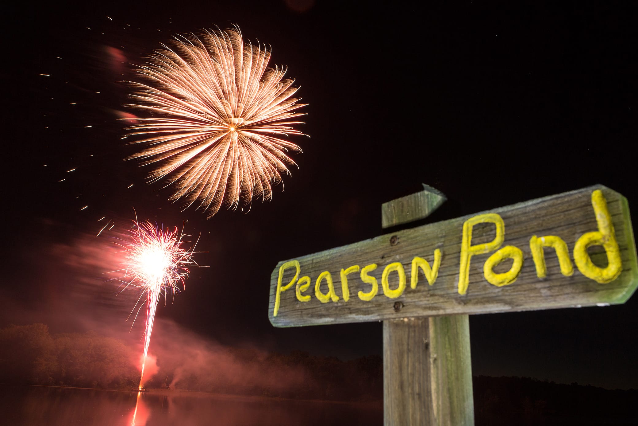 Fireworks burst in the night sky above a calm pond. In the foreground, a wooden sign with yellow letters reads Pearson Pond. Trees and water are visible in the background.