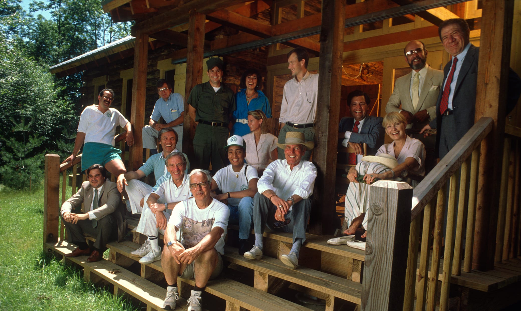 A group of people, casually dressed, sit and stand on the wooden steps of a rustic cabin surrounded by greenery, smiling and posing for a group photo.