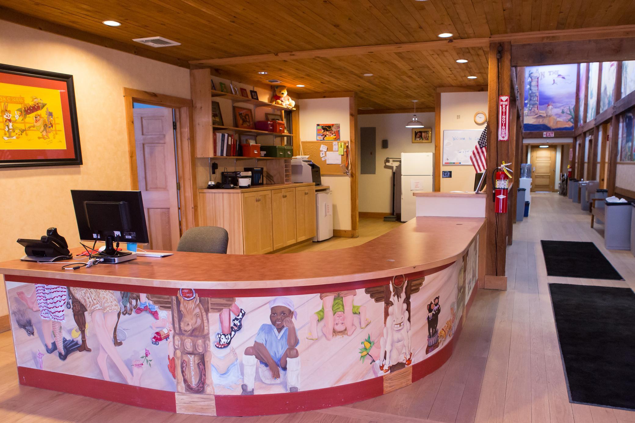 A welcoming reception desk with colorful painted art, computer, and office supplies. Wooden floors and ceiling, filing cabinets, and vibrant wall art create a warm, inviting office space.