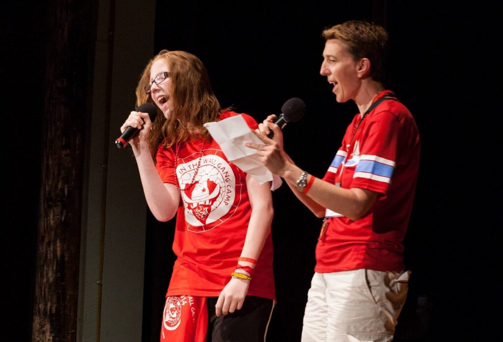 Two people in red shirts passionately perform on stage with microphones. One holds a paper while the other sings enthusiastically. They stand on a wooden floor with a dark background.