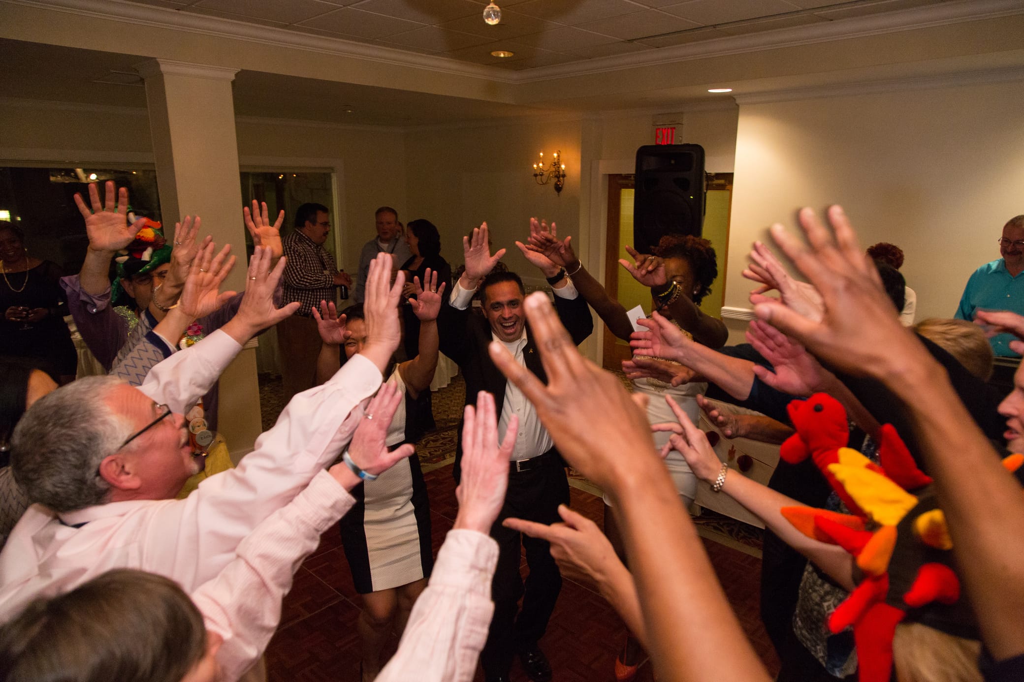 A group of people at an indoor party laugh and raise their hands in a circle, celebrating together. Some wear colorful hats. The room is warmly lit, and everyone looks joyful and engaged.