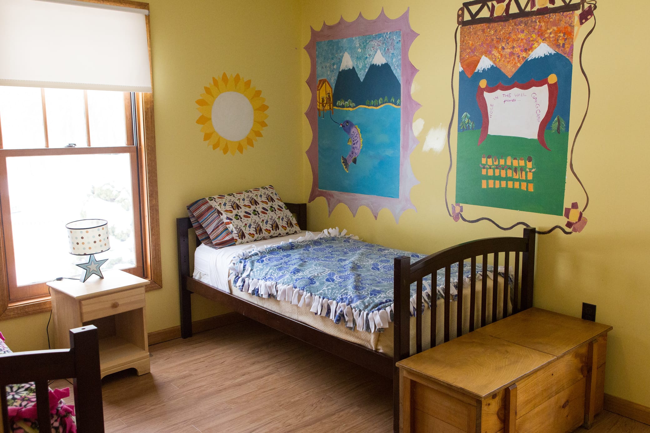 A cozy bedroom with a single bed, blue blanket, wooden furniture, and yellow walls decorated with colorful, fairy-tale themed murals and a sun painted beside a bright window.
