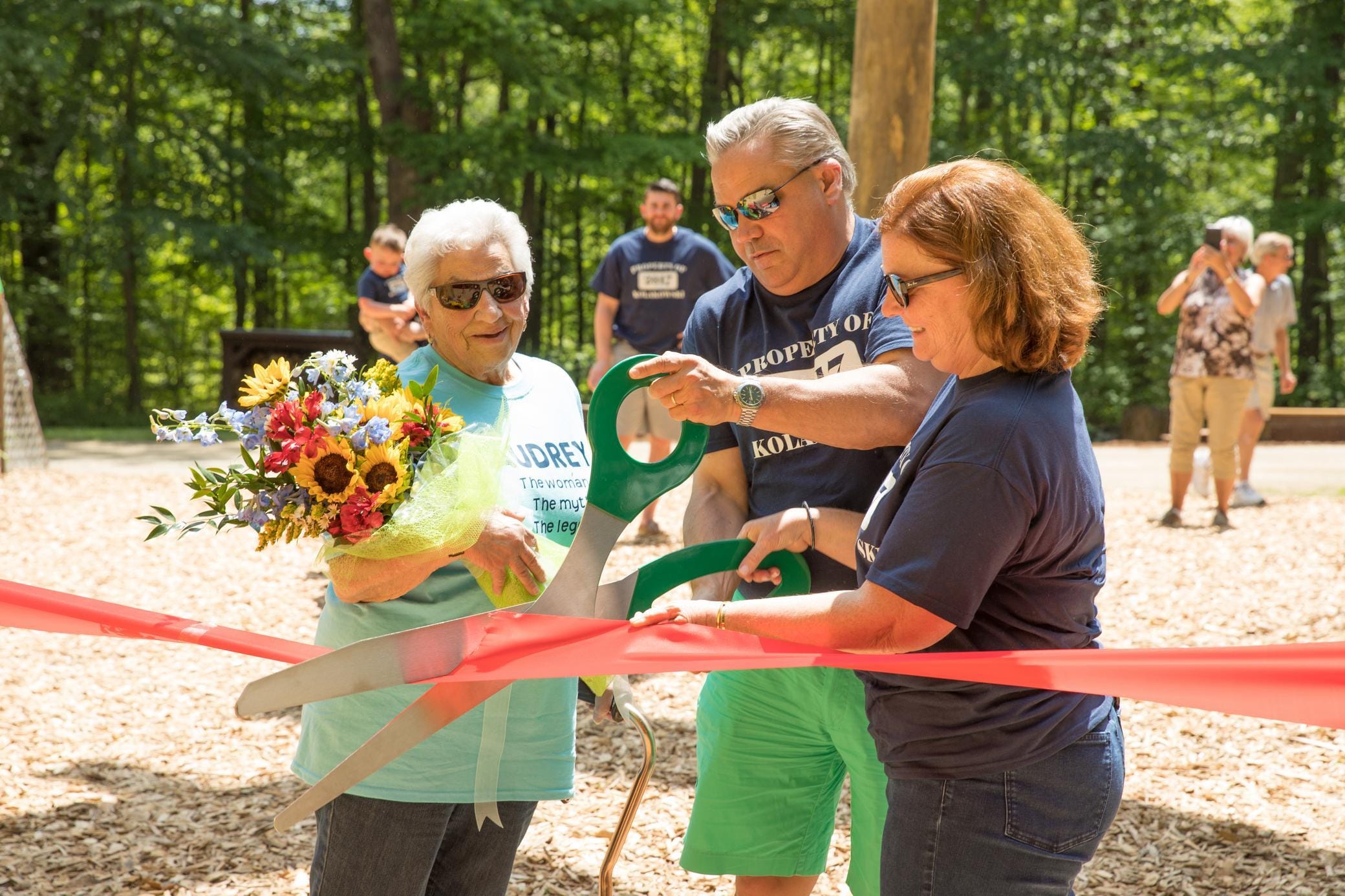 Three adults cut a red ribbon with oversized scissors at an outdoor event; one person holds a bouquet of flowers. Trees and a few other people are visible in the background.