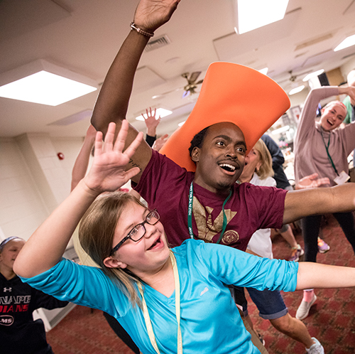 A man wearing an oversized orange hat and a girl in glasses smile and raise their arms while dancing in a lively indoor group activity. Other people are dancing in the background.