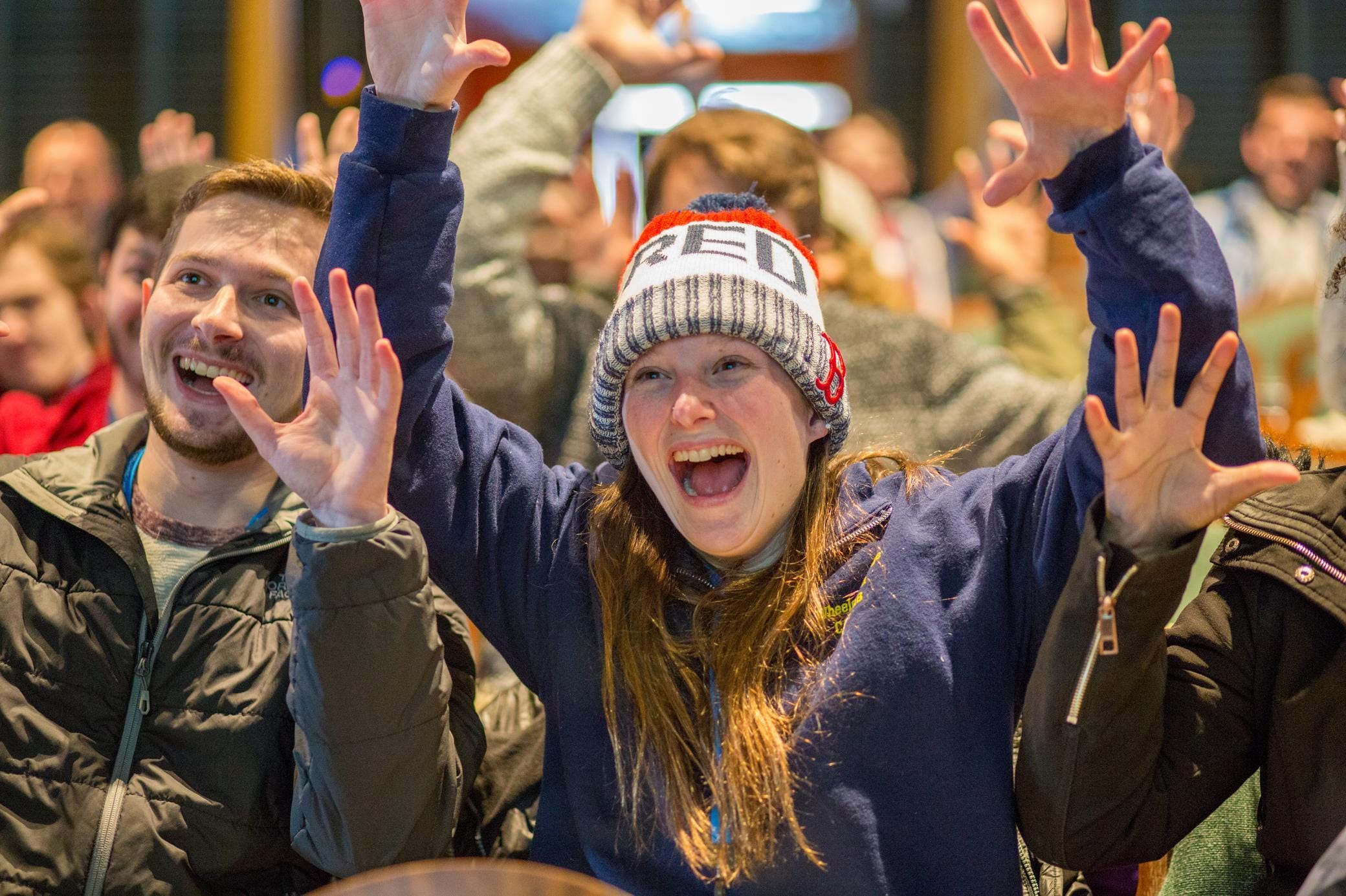 A group of people cheer enthusiastically indoors. A woman in front, wearing a beanie and blue sweater, raises her hands and smiles widely. Others around her also smile and raise their hands, showing excitement.
