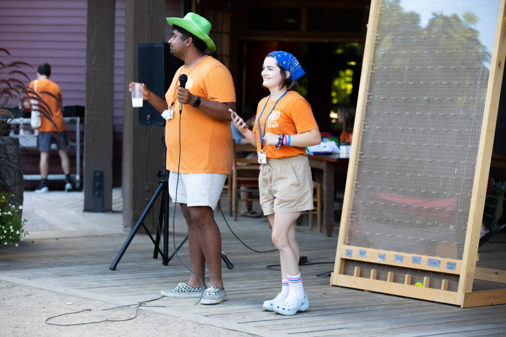 Two people in orange shirts stand on a wooden stage. One holds a microphone and a drink, the other holds a phone. They seem to be hosting an event. A large pegboard and others in orange shirts are visible in the background.