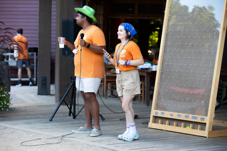 Two people in orange shirts stand on a wooden stage. One holds a microphone and a drink, the other holds a phone. They seem to be hosting an event. A large pegboard and others in orange shirts are visible in the background.