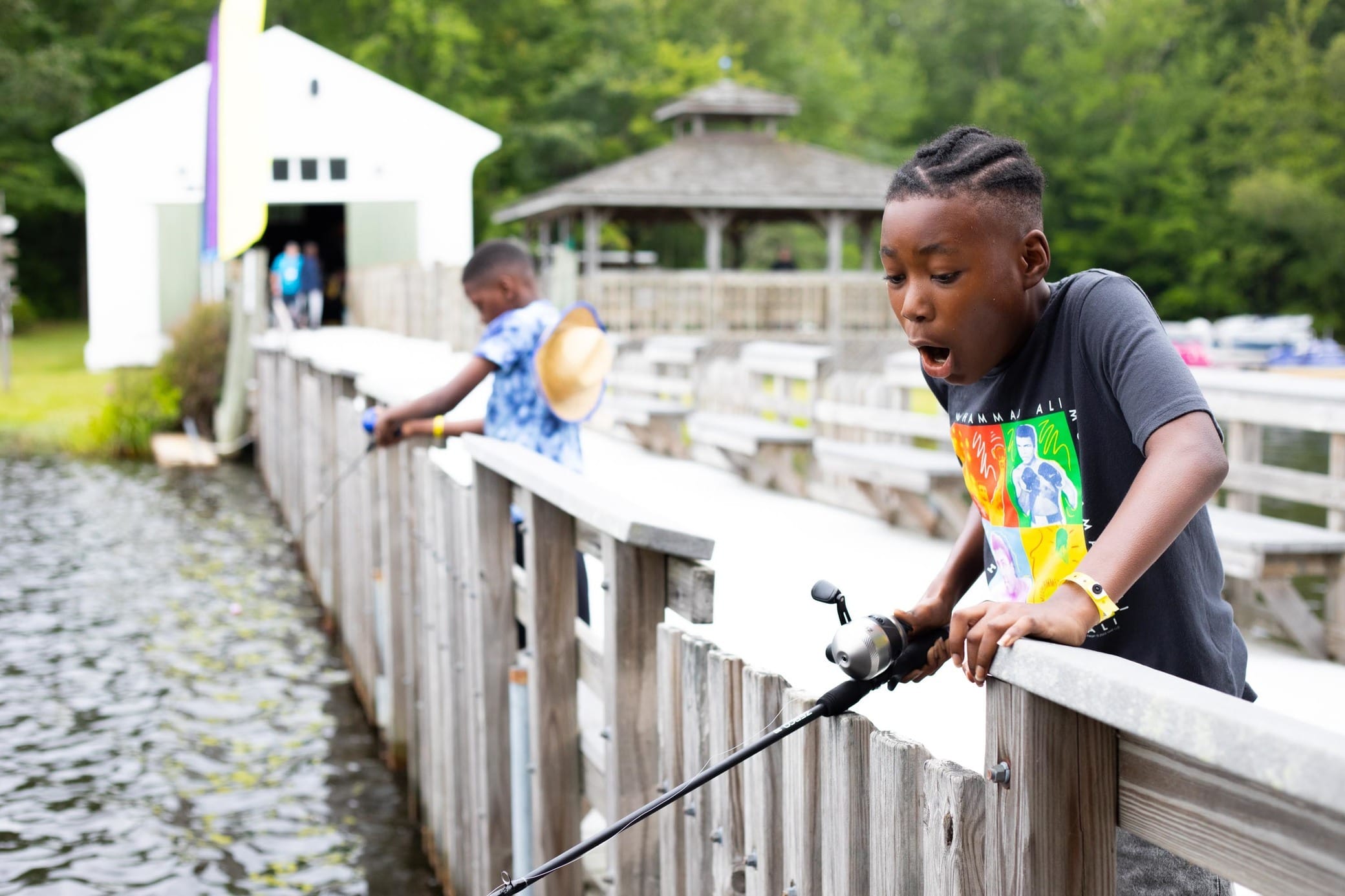 A surprised boy with braided hair holds a fishing rod over a wooden railing, looking into the water, while another child fishes in the background near a lakeside pier and gazebo.