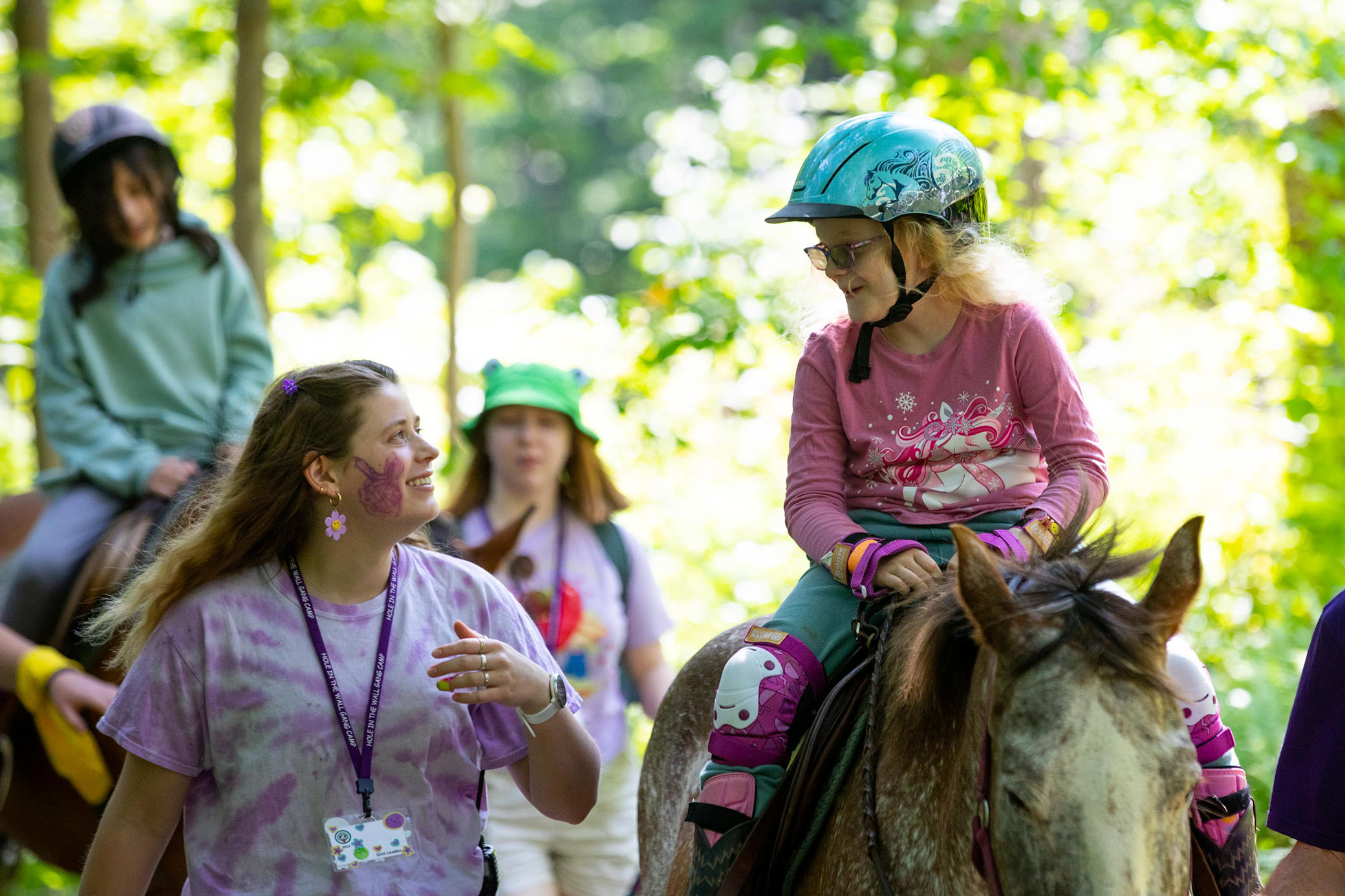 A girl wearing a helmet and pink shirt rides a horse along a wooded trail, smiling at a woman beside her. Other riders and counselors walk nearby in the background.