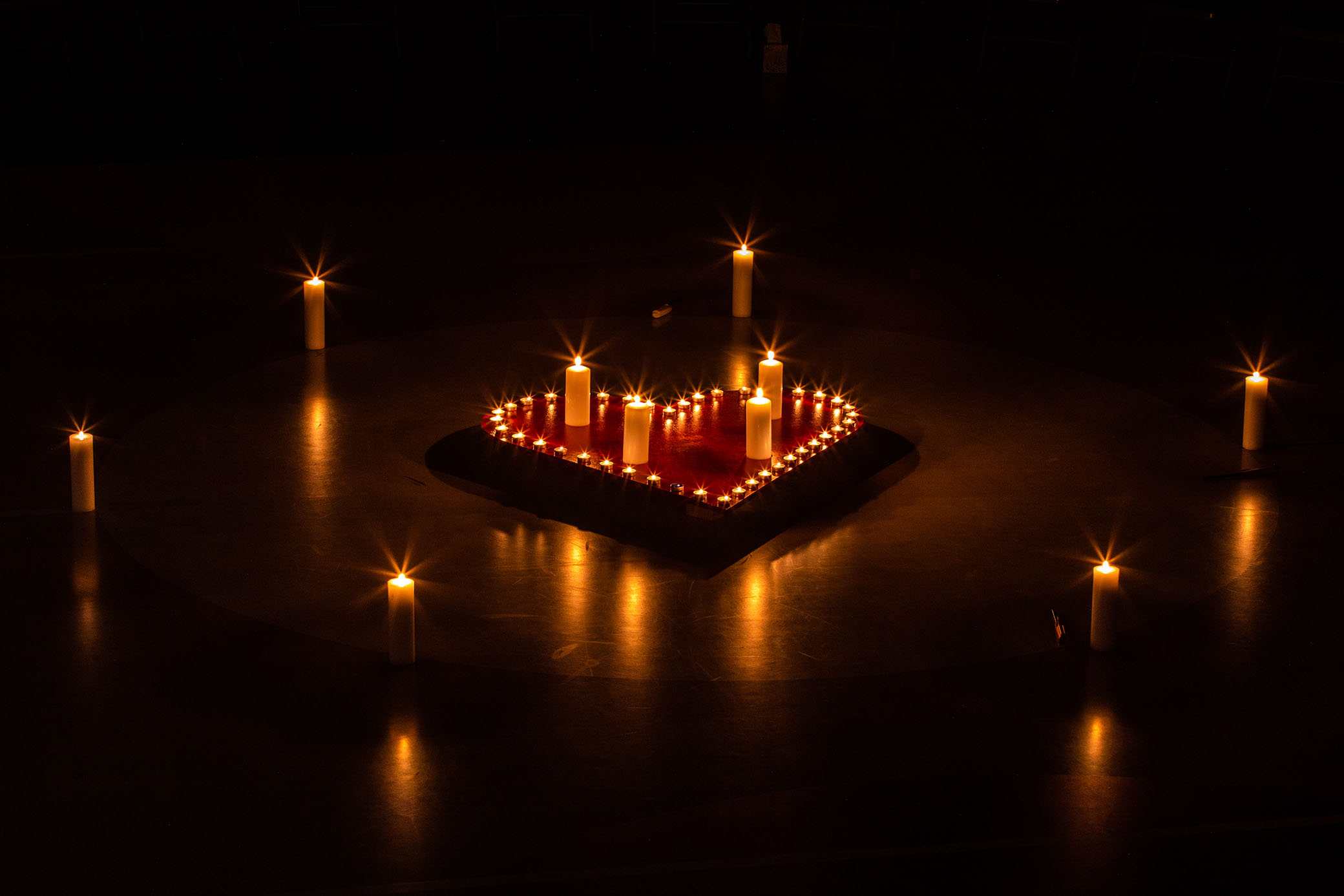 A heart shape made of small lit candles is surrounded by larger candles arranged in a circle on a dark surface, creating a warm, glowing ambiance.
