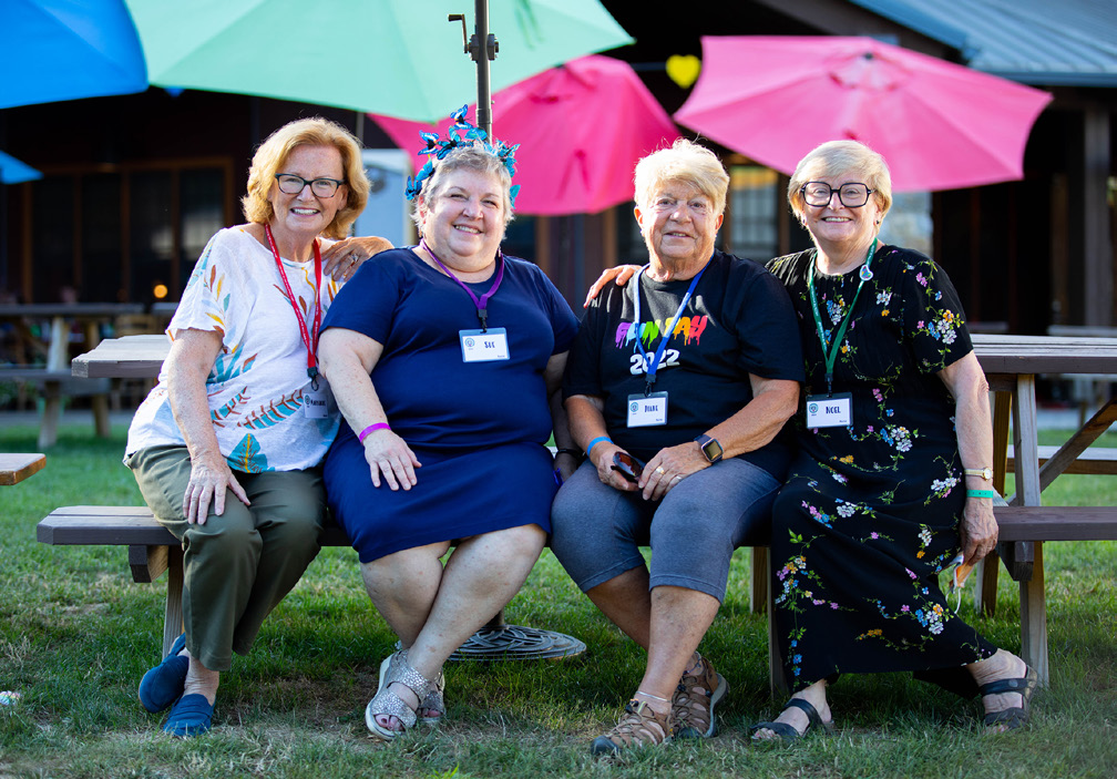 Four older women sit on a picnic bench outdoors, smiling at the camera. They wear name tags and colorful clothing, with umbrellas and a building in the background.