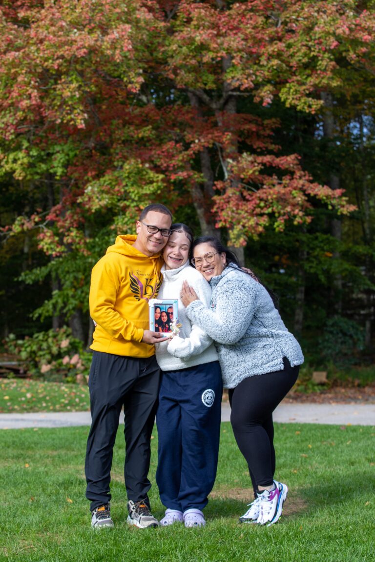 Three people smiling and hugging outdoors in front of autumn trees, with the person in the middle holding a framed family photo.
