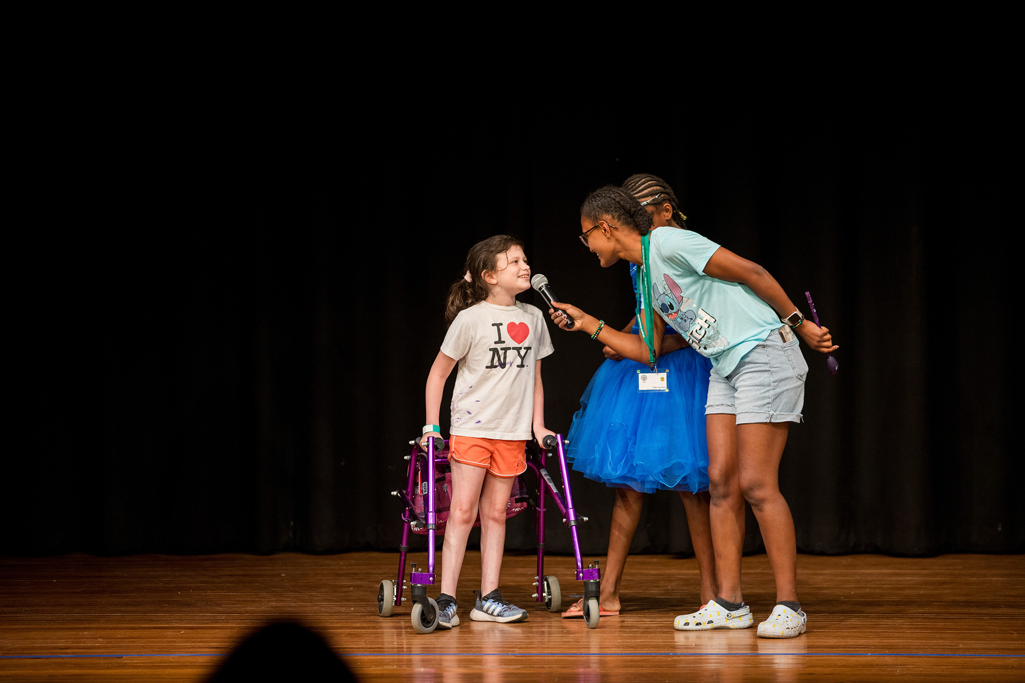 A young girl with a walker smiles as another girl holds a microphone to her on stage. They stand on a wooden floor with a black curtain in the background.