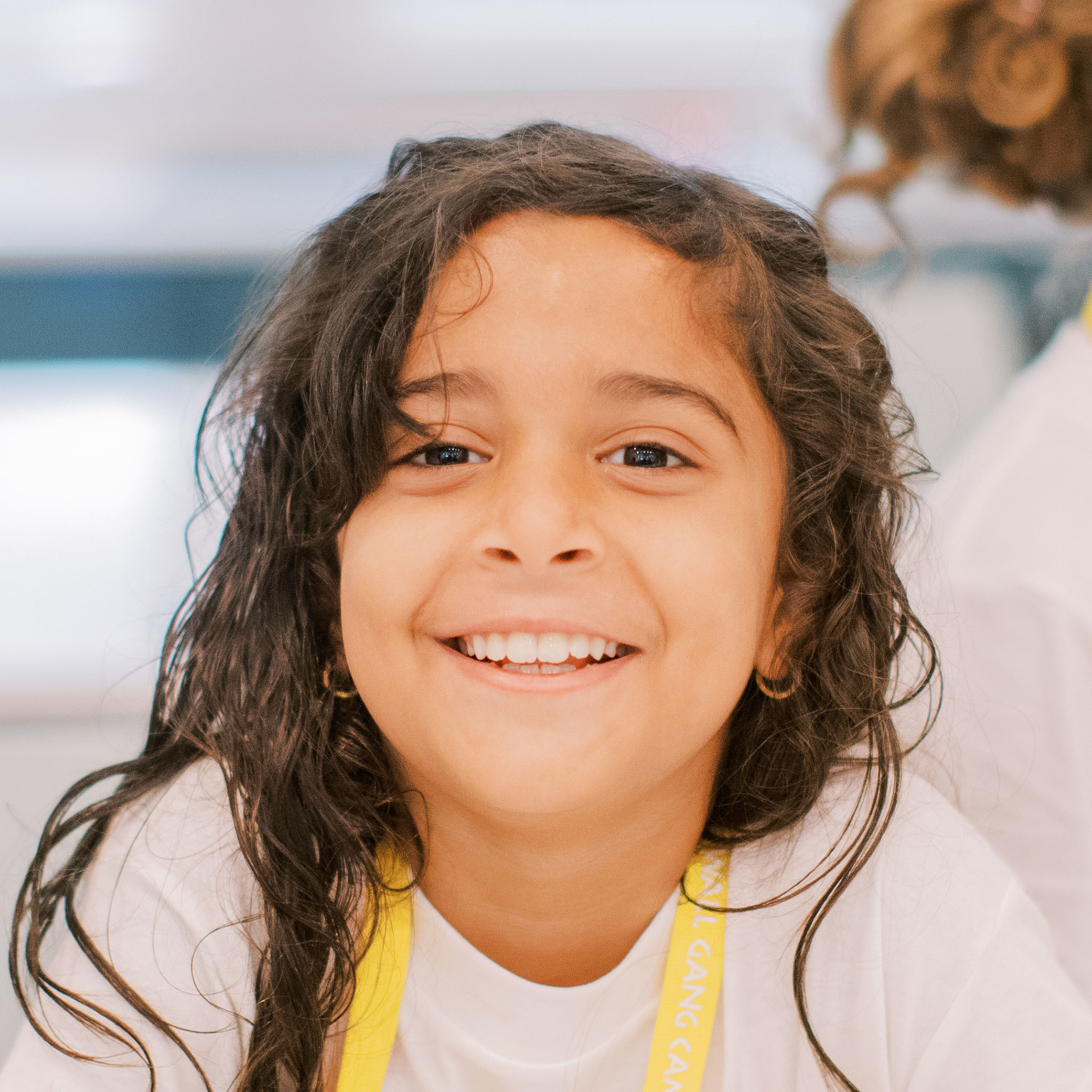A young girl with long dark hair smiles brightly at the camera, wearing a white shirt and a yellow lanyard. The background is softly blurred.