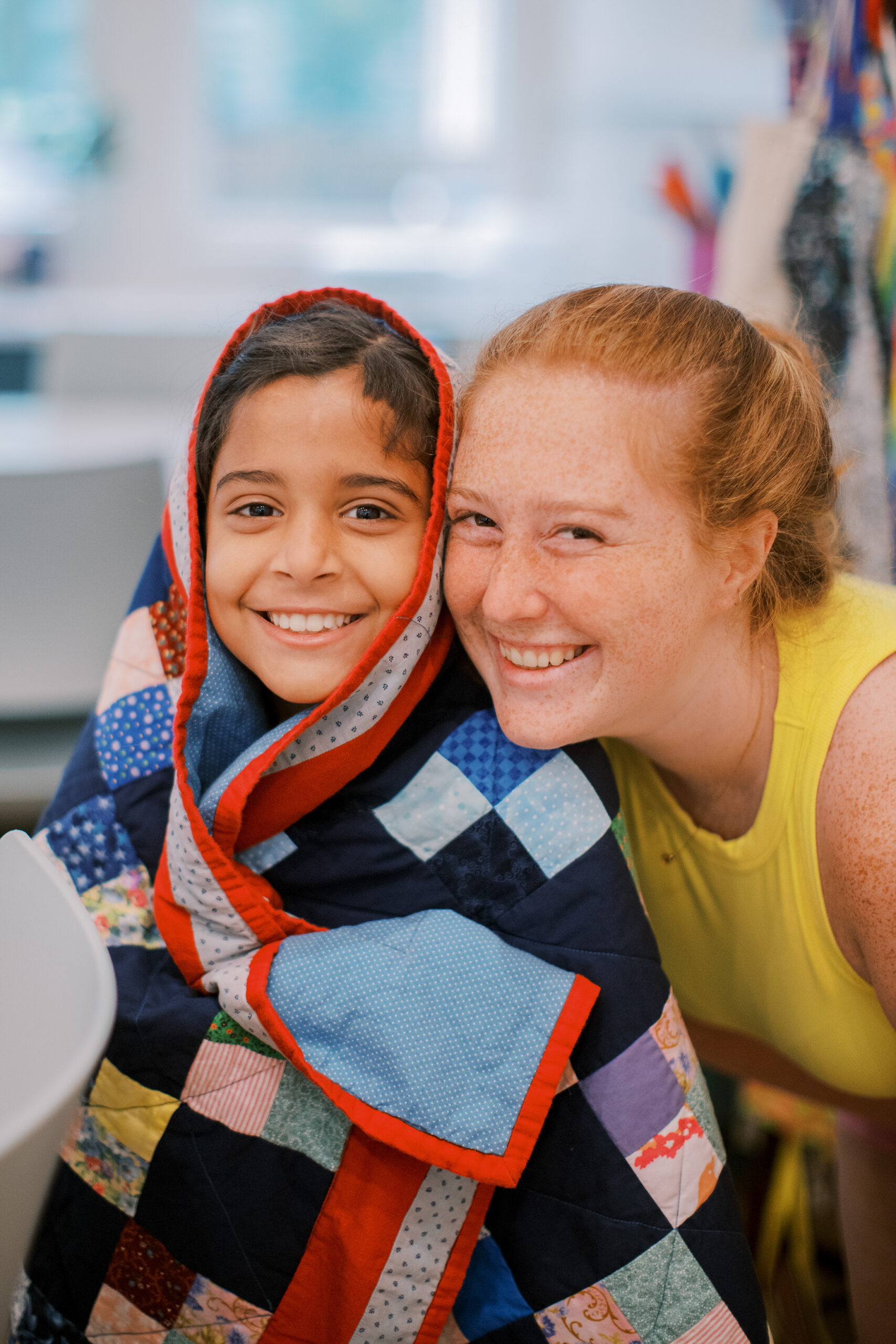 A smiling child wrapped in a colorful patchwork quilt sits next to a woman with red hair and freckles, both smiling at the camera in a bright, cheerful room.