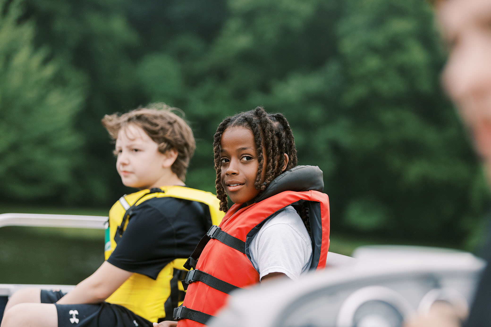 Two children wearing life jackets sit on a boat, facing forward. The child in front wears a red life jacket and looks at the camera, while the other wears yellow. Green trees are blurred in the background.