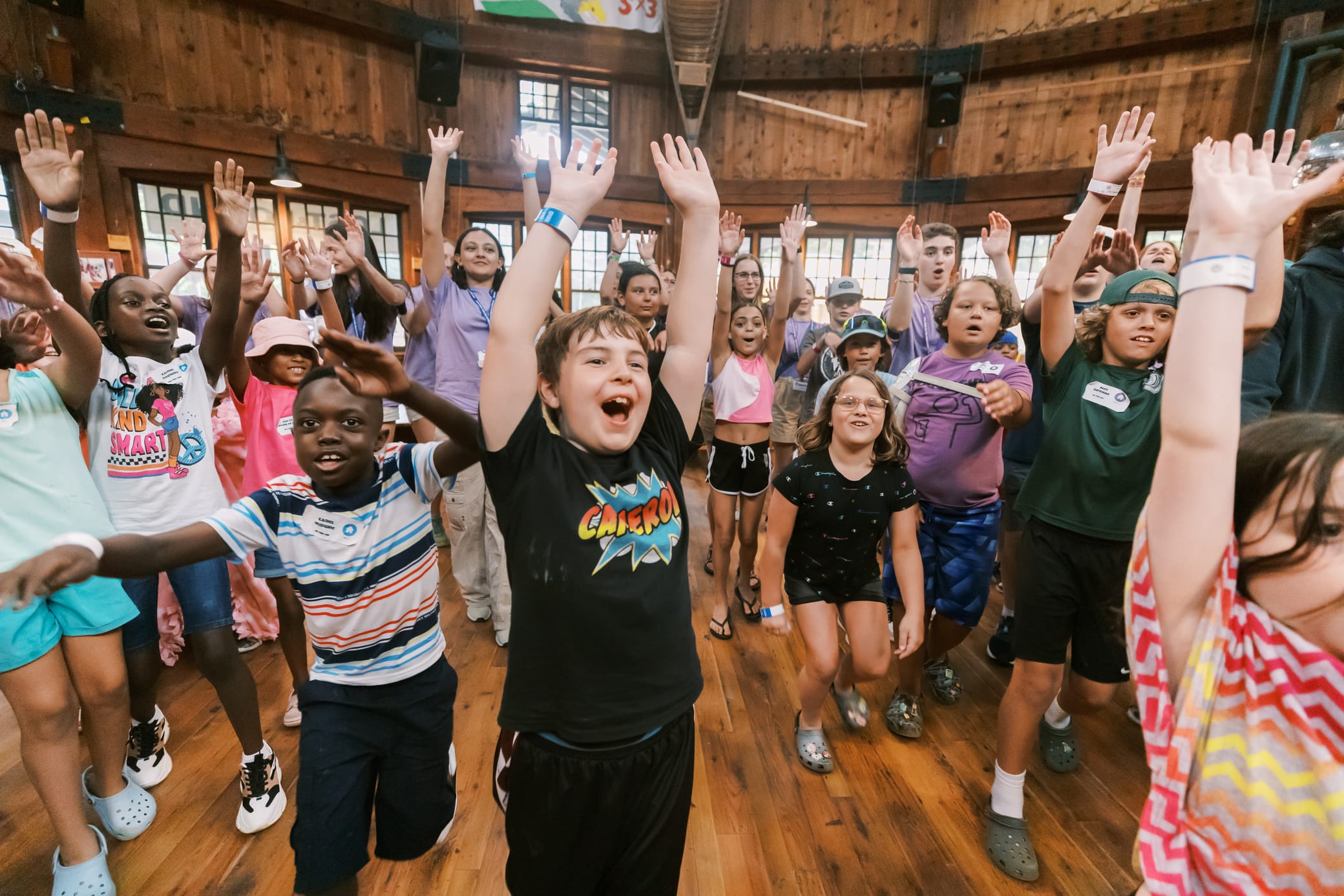 A group of children and adults cheerfully raise their hands in a wooden hall, smiling and laughing during an indoor activity or event.