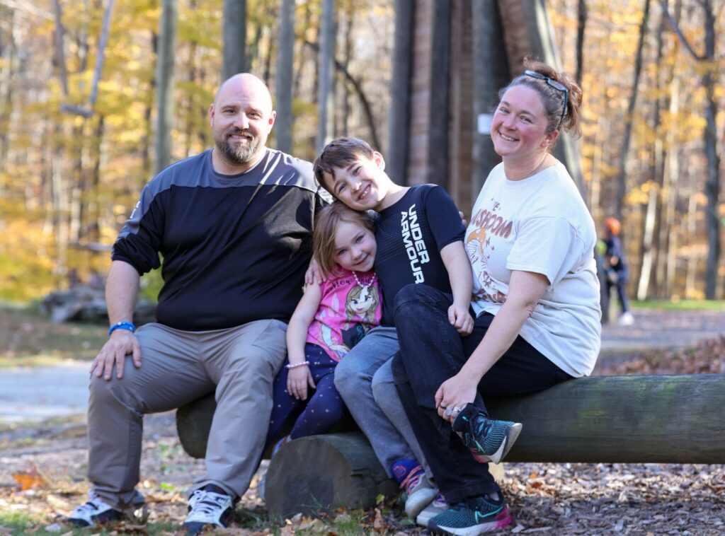 A smiling family of four sits on a log in a wooded park, with a tall ropes course and autumn trees in the background. Two adults and two children pose together, enjoying a sunny fall day outdoors.