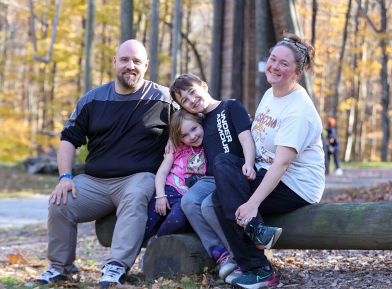 A smiling family of four sits on a log in a wooded park, with a tall ropes course and autumn trees in the background. Two adults and two children pose together, enjoying a sunny fall day outdoors.