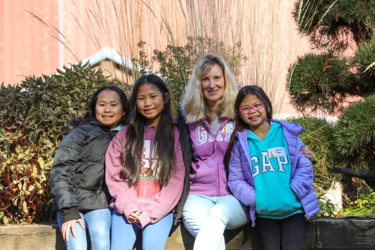 Four people, three children and one adult, sit together outdoors on a wooden bench, smiling at the camera. They wear hoodies, and green bushes and tall grasses are in the background.